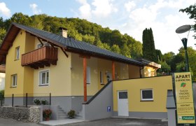Apartment Dirndltal, © Familie Krückel Gelbes Haus mit Balkon und Schild 'Apart Dirndltal' vor einem bewaldeten Hügel.
