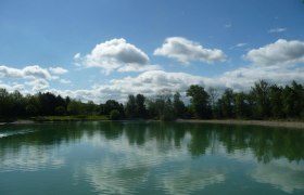 Ebersdorfer See, © zVg Gemeinde Ober-Grafendorf Ein ruhiger See mit Bäumen und Wolken, die sich im Wasser spiegeln.