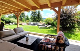 Außenbereich- Kräuterhäusl Puchenstuben, © Kräuterhäusl Puchenstuben Überdachte Terrasse mit Sitzmöbeln und Blick auf einen Garten mit Liegestühlen und Bäumen.