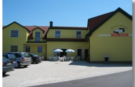 Pension Ziekel, © Pension Ziekel Gelbes Gebäude mit der Aufschrift 'Pension Ziekel', Autos und Tische mit Sonnenschirmen davor.