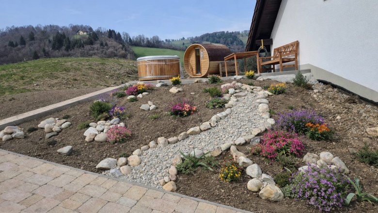 Haarstubenhäusl "Glückskäfer", © Sonja Filsner Ein gepflegter Garten mit Blumen, einem Kiesweg und einem Holzsauna-Fass neben einem Haus in ländlicher Umgebung.