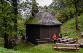 Hölzerne Kirche, © zur Verfügung gestellt: Gemeinde Schwarzenbach an der Pielach Eine kleine, hölzerne Kirche im Wald mit zwei Personen und Holzbänken im Vordergrund.