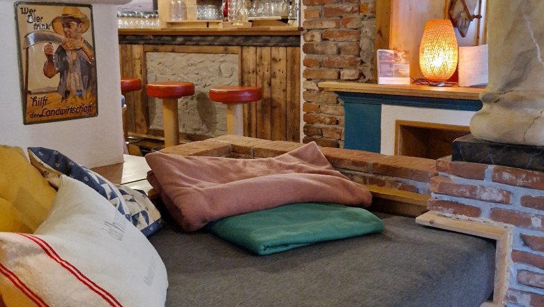 Aufenthaltsbereich- Kräuterhäusl Puchenstuben, © Kräuterhäusl Puchenstuben Gemütlicher Innenraum mit Holzmöbeln, Kissen und einer Bar im Hintergrund.