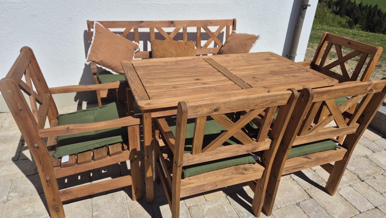 Haarstubenhäusl "Glückskäfer", © Sonja Filsner Holz-Gartenmöbel-Set mit Tisch, Stühlen und Bank auf einer Terrasse.