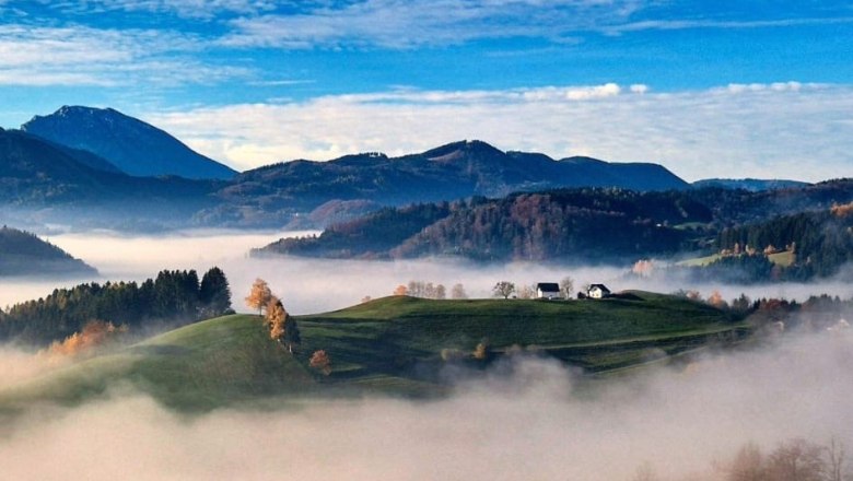 Haarstubenhäusl "Glückskäfer", © Sonja Filsner Eine malerische Landschaft mit grünen Hügeln, umgeben von Nebel und Bergen im Hintergrund unter einem blauen Himmel.