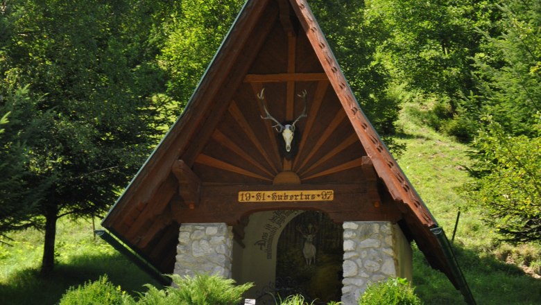 Hubertus-Kapelle, © Schwarzenbach/Piel. Eine kleine Kapelle mit spitzem Dach und Hirschgeweih, umgeben von Bäumen und Büschen.