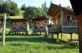 Erlebnisdorf Sulzbichl, © weinfranz.at Drei Holzhütten auf Stelzen mit Veranda in einem grünen, bewaldeten Gebiet.