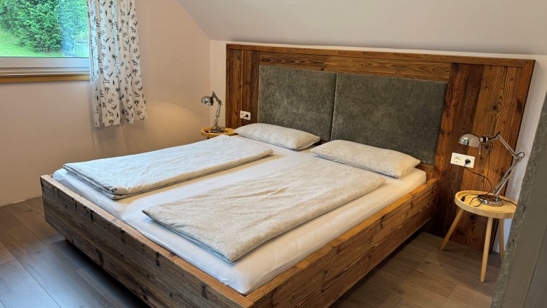 Einheit Turmkogel- Kräuterhäusl Puchenstuben, © Kräuterhäusl Puchenstuben Ein gemütliches Schlafzimmer mit einem Doppelbett aus Holz, zwei Nachttischen mit Lampen und einem Fenster mit Vorhängen.