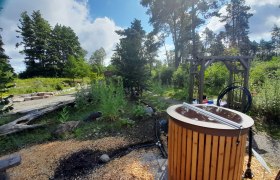 Regenwassernutzung, © © L.Riesenhuber Ein Garten mit einem Holzfass, umgeben von Bäumen und Pflanzen unter einem blauen Himmel.