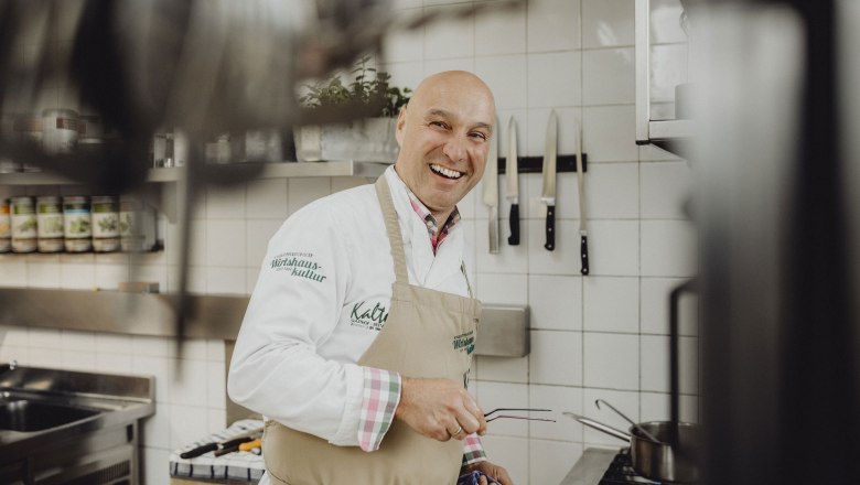 Ein lachender Koch in einer professionellen K&uuml;che mit Messern an der Wand.