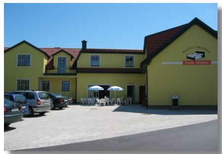 Pension Ziekel, © Pension Ziekel Gelbes Gebäude mit der Aufschrift 'Pension Ziekel', Autos und Tische mit Sonnenschirmen davor.