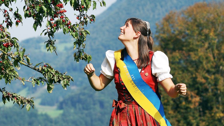 Frau in traditioneller Tracht steht unter einem Baum mit roten Früchten in einer ländlichen Landschaft.
