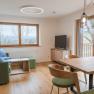 Modern eingerichtetes Wohnzimmer mit Holzboden, gr&uuml;ner Couch, Esstisch und gro&szlig;en Fenstern mit Blick ins Freie.