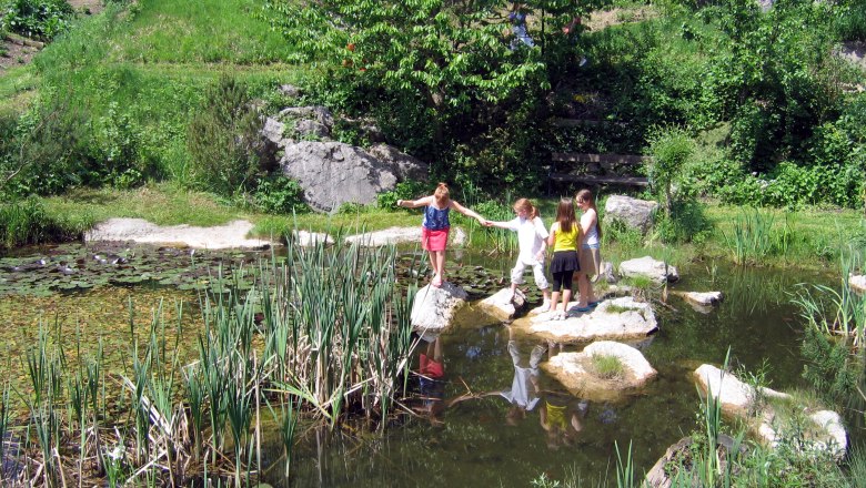Kinder spielen an einem Teich im Grünen mit Seerosen und Schilf.