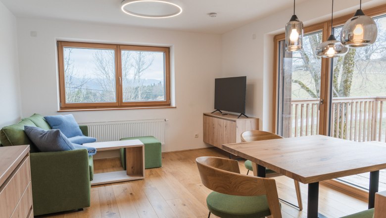 Modern eingerichtetes Wohnzimmer mit Holzboden, gr&uuml;ner Couch, Esstisch und gro&szlig;en Fenstern mit Blick ins Freie.