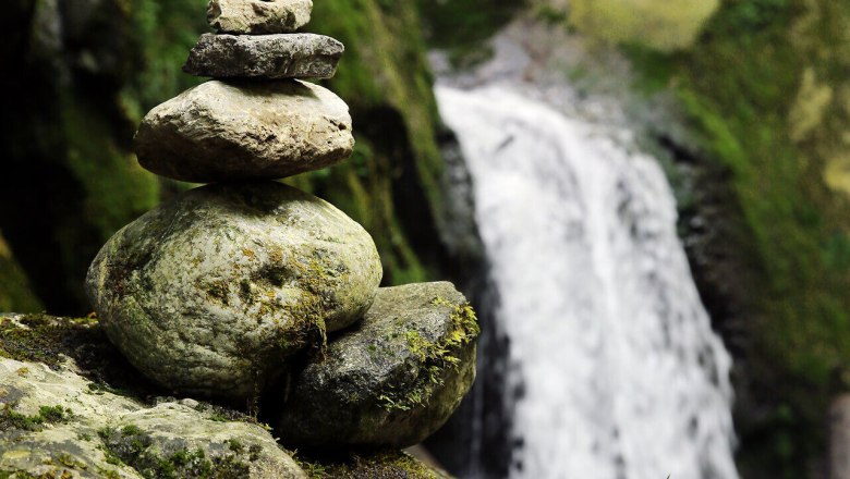 Sanfte Wasserf&auml;lle pl&auml;tschern inmitten einer idyllischen Landschaft, w&auml;hrend kunstvoll gestapelte Steine die Ruhe der Natur widerspiegeln. Umgeben von &uuml;ppigem Gr&uuml;n und dem sanften Rauschen des Wassers l&auml;dt dieser Ort zum Verweilen und Entspannen ein.