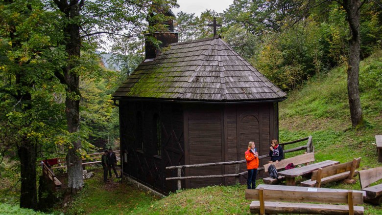 Eine kleine, h&ouml;lzerne Kirche im Wald mit zwei Personen und Holzb&auml;nken im Vordergrund.