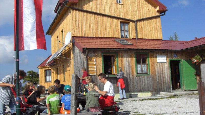 Eine Gruppe von Menschen sitzt vor einer Berghütte mit Holzfassade und österreichischer Flagge.