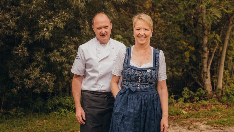 Ein Mann in Kochuniform und eine Frau in traditioneller Tracht stehen l&auml;chelnd vor einem Wald.