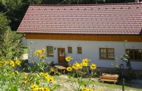 Ein weißes Haus mit rotem Dach, umgeben von gelben Blumen und grüner Landschaft.