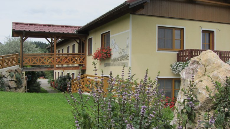 Ein gelbes Gebäude mit Holzbalkon und Blumen, umgeben von grüner Landschaft.
