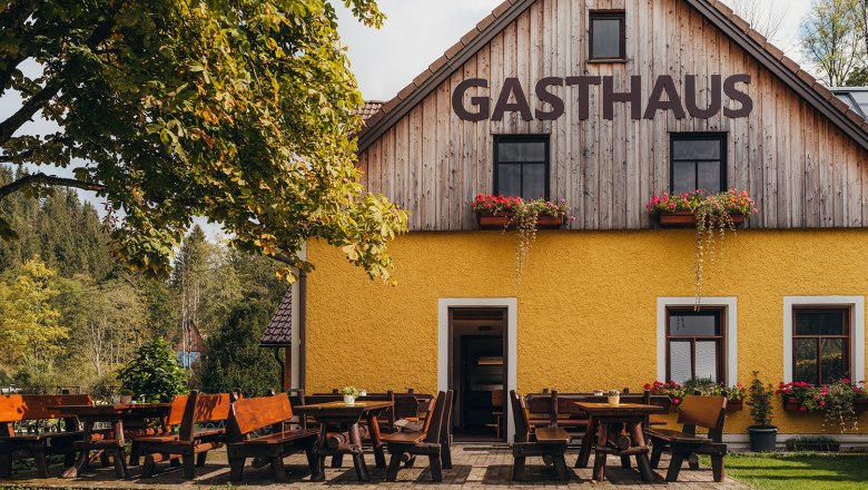 Ein gelbes Gasthaus mit Holztischen und B&auml;nken im Freien, umgeben von B&auml;umen und Natur.