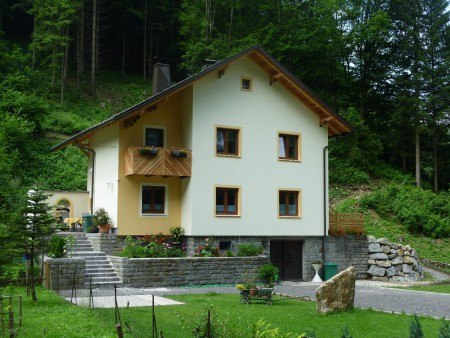 Ein gelbes G&auml;stehaus mit Holzbalkon, umgeben von gr&uuml;ner Natur und B&auml;umen.
