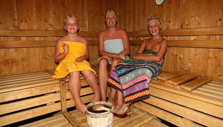 Drei Personen in einer Sauna, umgeben von Holzw&auml;nden, mit Handt&uuml;chern bekleidet.