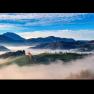 Eine malerische Landschaft mit gr&uuml;nen H&uuml;geln, umgeben von Nebel und Bergen im Hintergrund unter einem blauen Himmel.