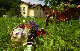 Ein gelbes Bauernhaus im Hintergrund, im Vordergrund ein Korb mit Gläsern und Flaschen auf einer Wiese.