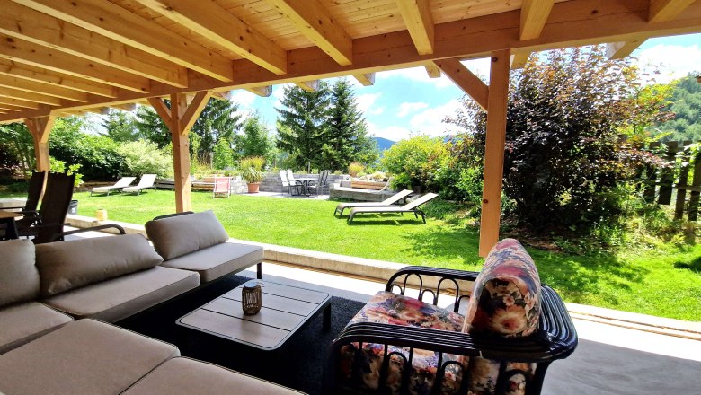 Außenbereich- Kräuterhäusl Puchenstuben, © Kräuterhäusl Puchenstuben Überdachte Terrasse mit Sitzmöbeln und Blick auf einen Garten mit Liegestühlen und Bäumen.