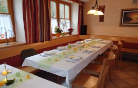 Ein festlich gedeckter Tisch in einem Gasthaus mit weißen Tischdecken, Kerzen und Blumen. Holzmöbel und Fenster mit Vorhängen im Hintergrund.