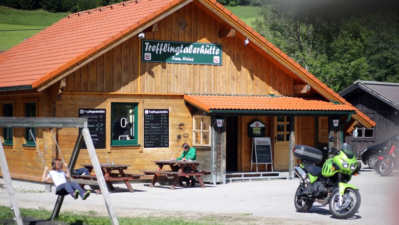 Eine Holzhütte mit dem Schild 'Trefflingtalerhütte', davor eine Schaukel, ein Motorrad und ein Mensch an einem Tisch.